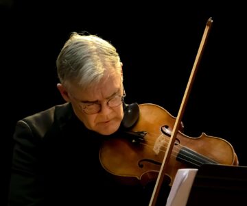 An older man with gray hair and glasses is playing a violin, focusing intently on his performance against a dark background.