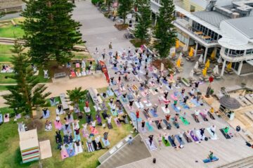 Aerial view of a large group practicing yoga on mats outdoors near a boardwalk, surrounded by trees and buildings, with a Ferris wheel and road in the background.