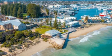 Aerial view of Fremantle Beach in Australia, with historic stone buildings, sandy shore, turquoise water, boats in the harbor, and green trees surrounding the coastal area on a sunny day.