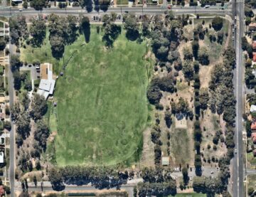 Aerial view of a large grassy oval sports field surrounded by trees, roads, and parking lots, with buildings and scattered cars visible along the edges.