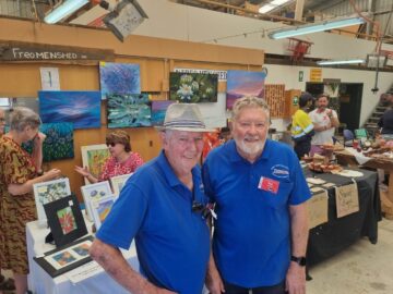 Two older men in blue shirts smile at a craft fair. Colorful paintings are displayed on the wall behind them. Other people browse art, talk, and look at cakes on a nearby table. The scene is indoors with a casual, welcoming atmosphere.
