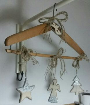 Wooden hanger with twine bows and three hanging ornaments: a star, an angel, and a tree, each with silver glitter accents. The hanger is suspended indoors near a white wall and light fixture.