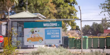 A sign on a chain-link fence reads Welcome to City of Fremantle Recycling Centre with illustrated recycling items. Colorful bins and a small building are visible behind the fence under sunny skies.