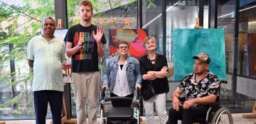 Five people with visible and invisible disabilities pose together indoors, smiling in front of colorful artwork; one uses a wheelchair, another a walker, and one a power wheelchair. Large windows and greenery are visible behind them.