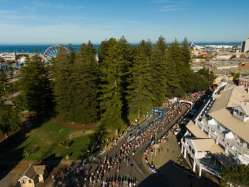 Aerial view of a large crowd gathered along a street lined with trees and buildings near a park, with a blue sky, ferris wheel, and ocean visible in the background.