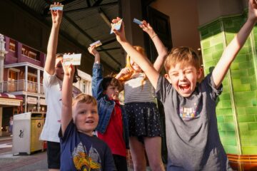 Five excited children stand outside, holding up tickets and smiling in the sun. One boy in front is cheering with arms raised, while other kids grin and pose near a green-tiled building on a city street.