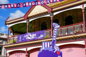 A Fremantle Dockers banner and decorations are displayed on a historic building, with a cartoon giraffe figure wearing purple, celebrating the team. The sky is bright and blue.