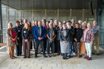A diverse group of about 25 adults stands together indoors, posing and smiling for a group photo. The background features glass walls and a modern metal grid.