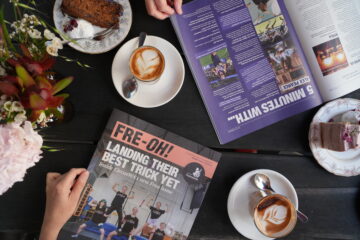 A table with coffee cups, cake slices, flowers, and two magazines, one open and one closed. The closed magazine cover reads, FRE·OH! Landing their best trick yet. Two peoples hands are visible.
