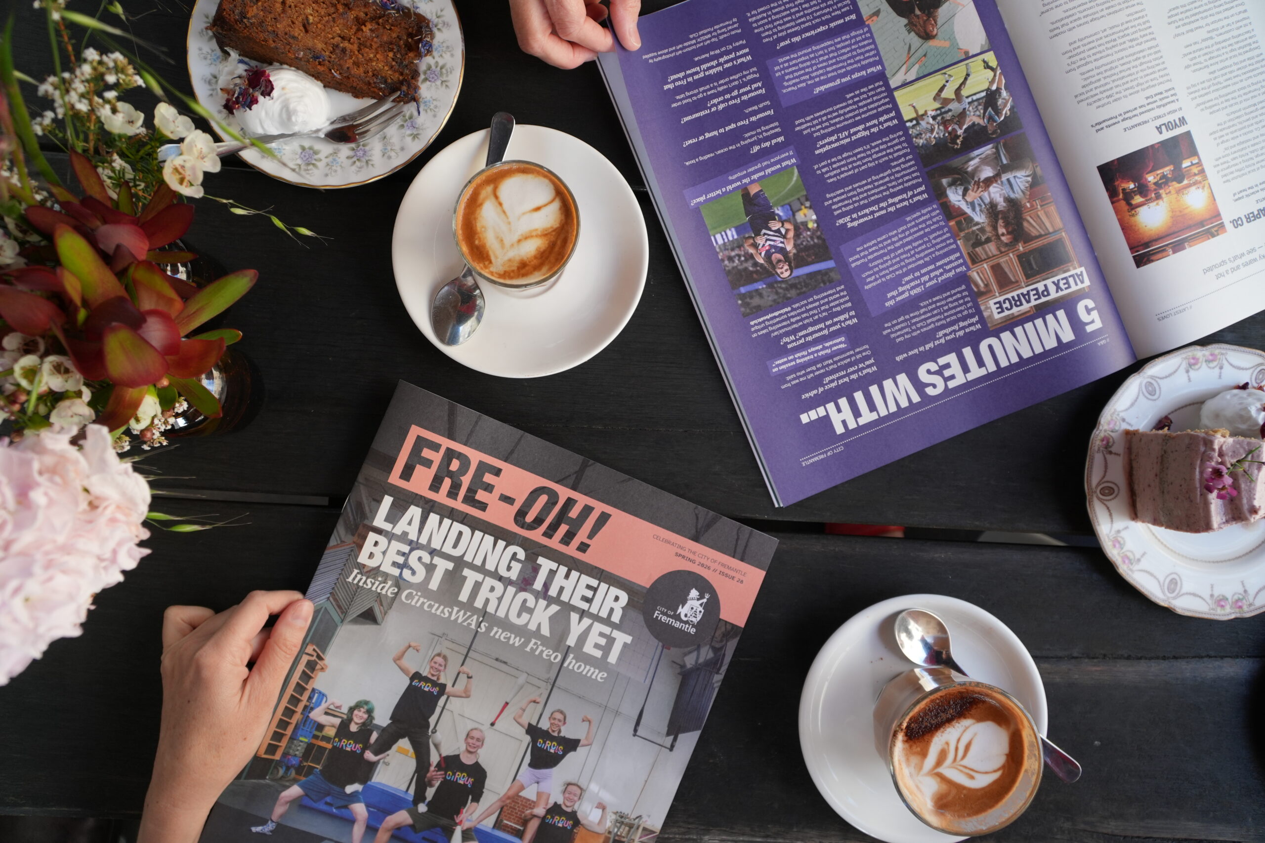 A table with coffee cups, cake slices, flowers, and two magazines, one open and one closed. The closed magazine cover reads, FRE·OH! Landing their best trick yet. Two peoples hands are visible.