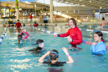 A swim instructor in a red shirt smiles and gestures while teaching a group of children in an indoor pool. The kids wear goggles and swim gear, practicing in a lane, with another instructor nearby.