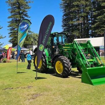 A green John Deere tractor with a front loader is displayed on grass at an outdoor event, with banners, trees, and people in the background under a blue sky.