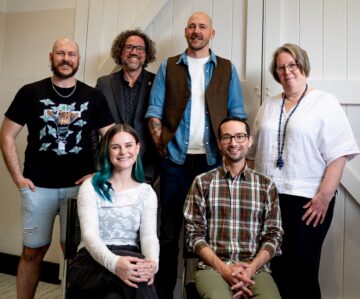 Six people pose indoors, with three seated in front and three standing behind them. They are casually dressed and smiling, in a bright room with white paneled walls and a door.