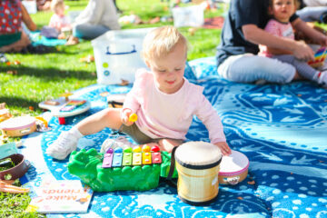 A toddler sits on a blue picnic blanket outdoors, playing with a drum, a xylophone, and a tambourine. Other children and adults are visible in the background. Sunlight brightens the scene.