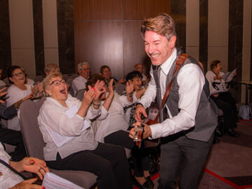 A smiling musician with a guitar performs for a group of elderly people seated in chairs; some are clapping and laughing, creating a joyful atmosphere at an indoor event.
