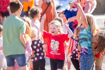 A joyful group of children play outdoors. One child in a red Merry Christmas shirt smiles with arms raised while others cheer and laugh around them in bright sunlight.