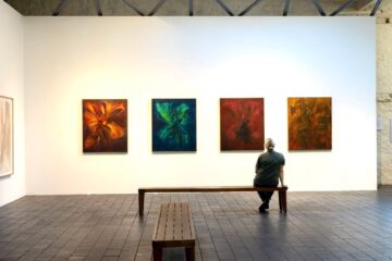 A person sits on a wooden bench facing four colorful abstract paintings on a white gallery wall under exposed beams.