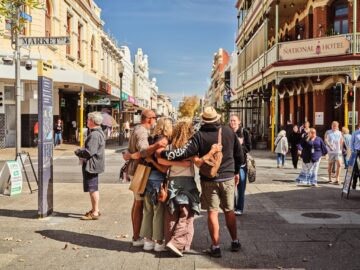 A group of people stand in a cheerful embrace in the middle of a busy city street, surrounded by historic buildings, shops, and pedestrians on a sunny day.