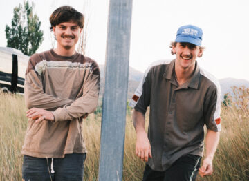 Two young men stand in tall grass, one leaning against a metal pole and the other smiling with a hand on the pole. Both wear casual clothes; one has a blue cap. Trees and hills are visible in the background.