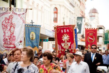 A crowd walks down a city street during a parade, holding colorful religious and cultural banners. People of various ages participate, with historic buildings in the background.
