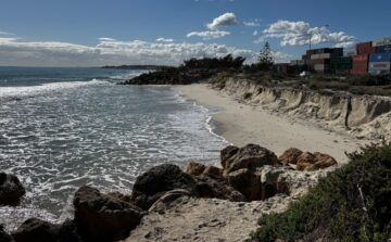 A sandy beach with gentle waves, rocky foreground, and a line of shipping containers stacked near the shore under a blue sky with scattered clouds.