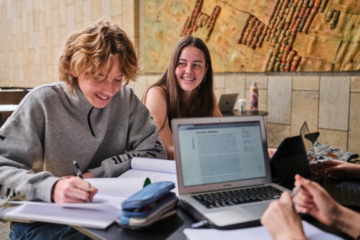 Two students sit at a table, studying and smiling. One writes in a notebook while the other looks on. Open laptops and study materials are on the table, with a map or artwork visible on the wall behind them.