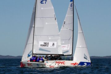 Two white sailboats with crews in blue shirts racing on the water. The sails display signs, including THIS IS FREMANTLE. The sky is clear, and distant land is visible on the horizon.