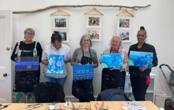 Five women stand indoors, each holding a blue-themed painting. Art supplies cover the table in front of them. Three framed photo collages and a decorative branch hang on the white wall behind them.