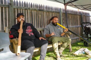 Two men sit on a wooden bench outdoors, each playing a didgeridoo. One instrument is wooden, the other yellow. They are under a canopy near a wooden fence, with sunlight casting shadows on the grass.