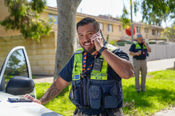 A smiling police officer stands outdoors by a car, talking on a cell phone and writing on a clipboard. Another officer is in the background, also using a phone. Residential buildings and trees are visible.