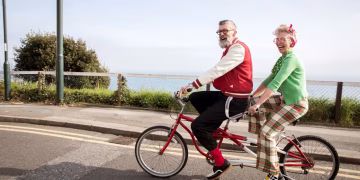 Two smiling adults ride a red tandem bicycle along a seaside road. The man wears a red jacket; the woman wears a green top and plaid pants. Both look cheerful and are enjoying the day.