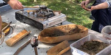 People use a portable gas stove and various wooden tools to work with clay and natural materials outdoors on a sunny day, possibly for a craft or educational activity.