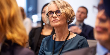 An older woman with short gray hair and round glasses attentively listens during a conference, surrounded by other seated attendees in business attire.