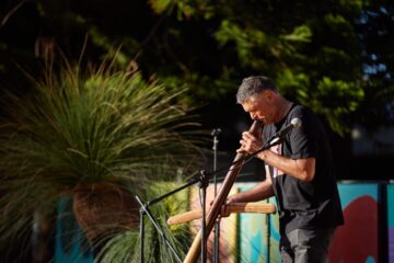 A man playing a didgeridoo into a microphone on an outdoor stage.