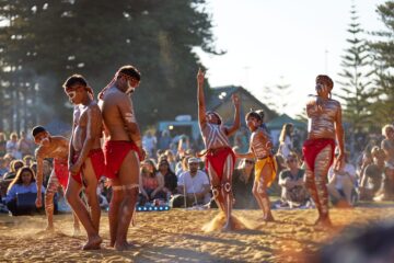 A group of Indigenous Australian men and boys performing a traditional dance on a beach, with body paint and red loincloths, in front of a seated audience.