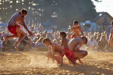 Indigenous performers wearing red loincloths and body paint dance and crouch on a sandy field before a seated audience.
