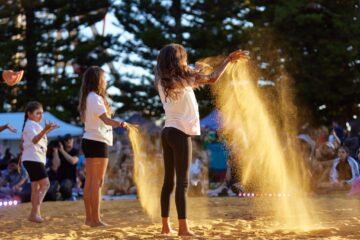 Two girls standing on a beach throwing yellow powder into the air.