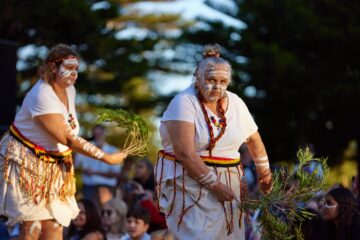 Two Indigenous women perform a traditional dance while wearing white face paint, necklaces, and skirts adorned with colorful tassels, each holding a bundle of eucalyptus leaves.