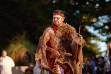 An Indigenous Australian man with white body paint wearing a fur cloak and a headband holds a boomerang during a performance.