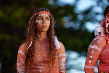 A young Indigenous woman with long curly hair wearing a traditional headband and white ochre markings on her face and arms.