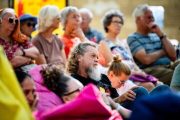 An audience of diverse adults sitting outdoors, focused on an off-screen event.