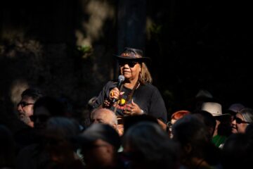 A woman wearing a wide-brimmed hat and sunglasses speaks into a microphone before a crowd.