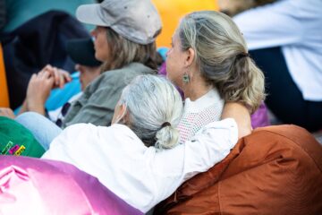A woman with gray hair resting her head on the shoulder of another woman, both seated among a group of people.