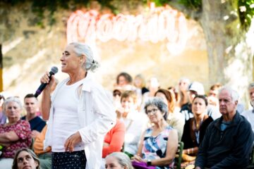 A woman with gray hair speaking into a microphone in front of an audience with a neon sign in the background.