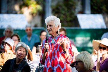 An elderly woman with short white hair, wearing a vibrant red and blue patterned tunic and a large turquoise necklace, speaks into a microphone before an audience.