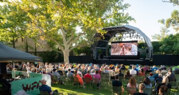 An outdoor audience sits on chairs and beanbags in a park, watching a movie projected onto a large screen under a stage canopy.
