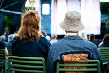 Two people sitting in green chairs seen from behind, watching a performance on an outdoor screen.