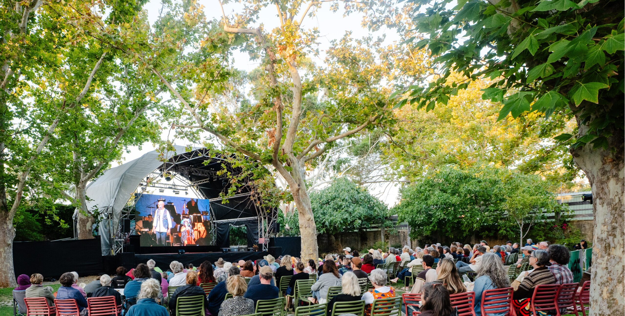 An audience sits in outdoor chairs watching a performance on a large screen at an open-air stage shaded by large trees.