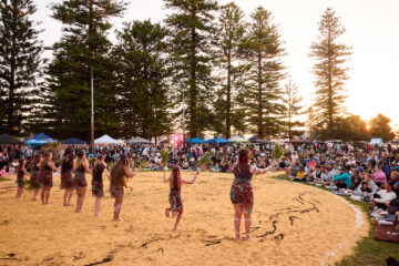 A group of Indigenous performers in traditional clothing dance in a circle on a sandy beach for a large seated audience gathered among pine trees at sunset.