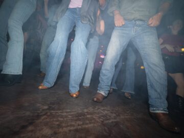 People in jeans and cowboy boots dancing on a wooden floor in a dimly lit bar.